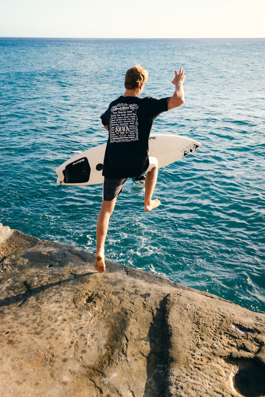 AVVA ambassador jumping off a rocky ledge into the ocean with a surfboard