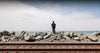 A person wearing a black shirt and green pants stands on large gray rocks near the ocean, with railroad tracks in the foreground and a cloudy sky above.