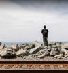 A person wearing a black shirt and green pants stands on large gray rocks near the ocean, with railroad tracks in the foreground and a cloudy sky above.