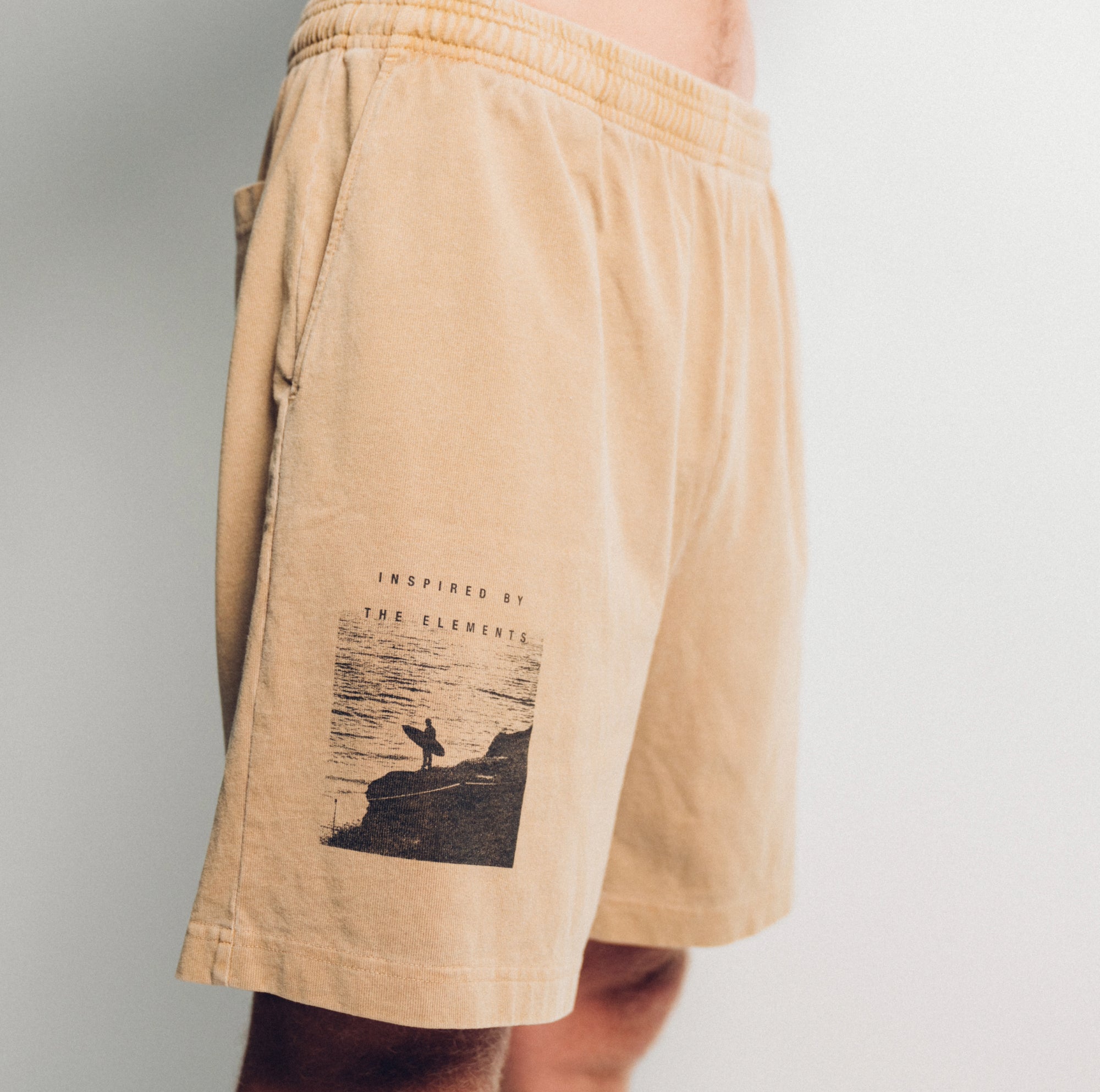 SIDE CLOSE UP VIEW OF KHAKI PIGMENT DYED HILO SHORT INSPIRED BY THE ELEMENTS LOGO WITH GRAPHIC OF SURFER HOLDING BOARD LOOKING AT WATER.