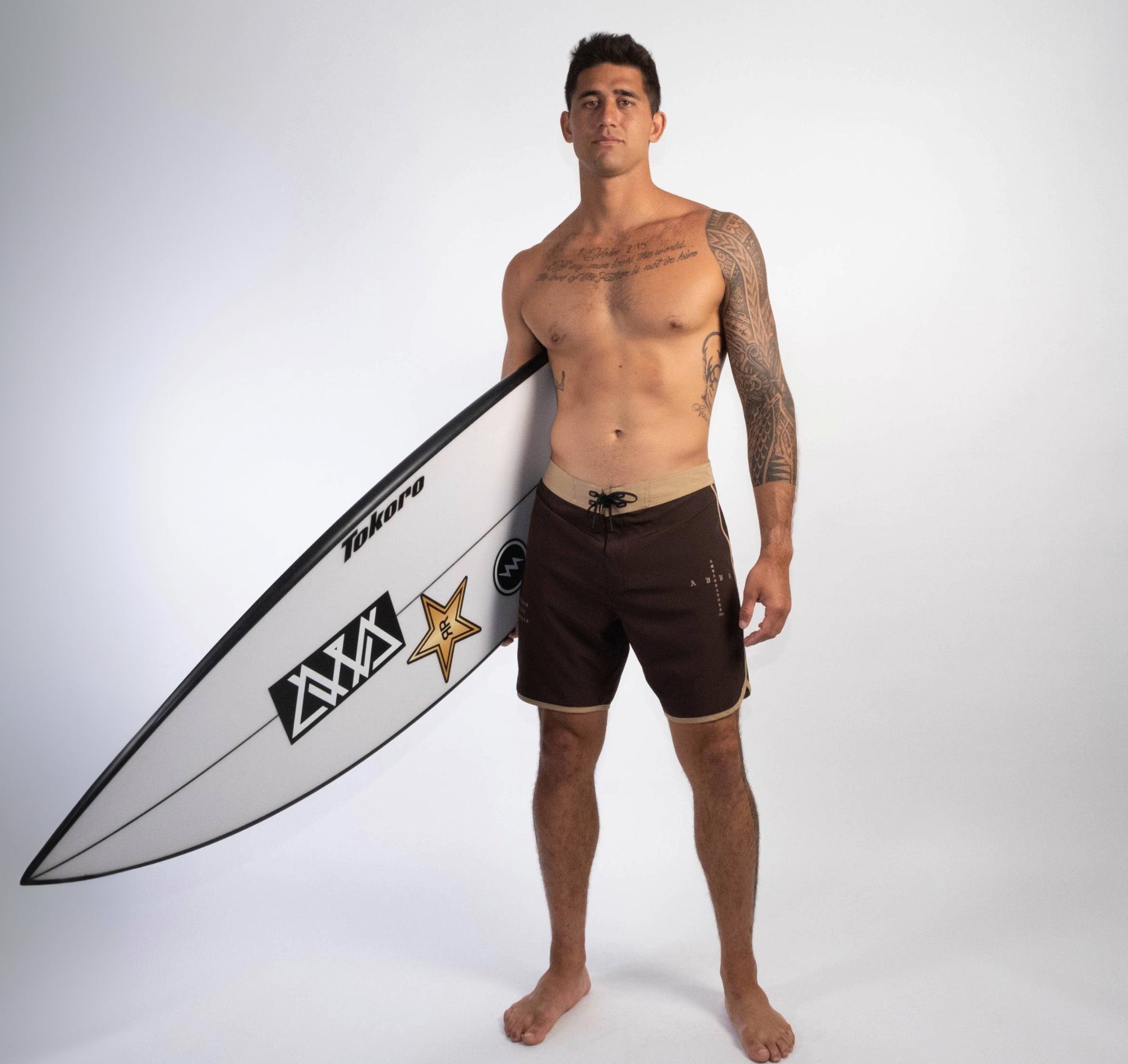 FRONT VIEW OF BROWN WEST SHORE AMBASSADOR BOARDSHORT WORN BY MODEL HOLDING A SURFBOARD.