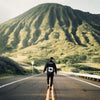 MODEL WALKING IN MIDDLE OF ROAD TWOARD GREEN MOUNTAIN IN HAWAII. WEARING AUMAKUA HOODIE. LARGE SCREEN PRINT ON BACK.