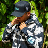 QUARTER VIEW OF MODEL WEARING BRODY HOODIE WITH TREE BACKGROUND. DETAILED VIEW OF SLEEVE SCREEN PRINT AVVA BLOCK LOGO. MODEL IS ALSO WEARING KA IWI HAT