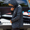 SIDE VIEW OF MODEL WEARING HANA ROAD ANORAK BLACK CAMO WINDBREAKER. HE IS ZIPPING UP THE JACKET WITH THE TRUCK IN BACKGROUND.