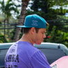 SIDE VIEW OF MODEL, KEKOA, WEARING REPRESENT SHARK TEAL HAT. EMBROIDERED INSPIRED BY THE ELEMENTS ON SIDE LIGHT LAVENDER .
