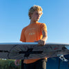 FRONT VIEW OF MODEL WEARING COUNTY FADED ORANGE PIGMENT TEE WITH HIS SURFBOARD.