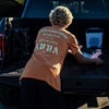 BACK VIEW OF MODEL WEARING COUNTY FADED ORANGE PIGMENT TEE PULLING HIS SURF BOARD OUT OF THE TRUCK. LARGE DISTRESSED SCREEN PRINT AMBASSADOR INSPIRED BY THE ELEMENTS AVVA LOGO ON BACK.