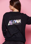 BACK VIEW OF FEMALE MODEL WEARING BOLT BLACK LONG SLEEVE. LARGE SCREEN PRINT REPEAT ALOHA OUTLINE PRINT. ONE HOLOGRAPHIC ALOHA
