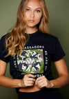 FRONT VIEW OF FEMALE MODEL WEARING COAST TO COAST BLACK TEE. CAMO PRINT DETAIL WITH AVVA LOGO. WE ARE THE AMBASSADORS. IN STUDIO WITH GREEN BACKGROUND.