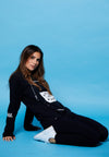 QUARTER FRONT VIEW OF FEMALE MODEL WEARING DIESEL BLACK LONG SLEEVE IN STUDIO WITH BLUE BACKDROP. LARGE AVVA LOGO SCREEN PRINT DETAIL ON FRONT. SMALL AVVA LOGO ON SLEEVE. FRONT PRINT WE ARE THE AMBASSADORS WITH ALOHA