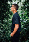 SIDE VIEW OF MODEL WEARING NIXON BLACK TEE WITH GREENERY IN BACKGROUND